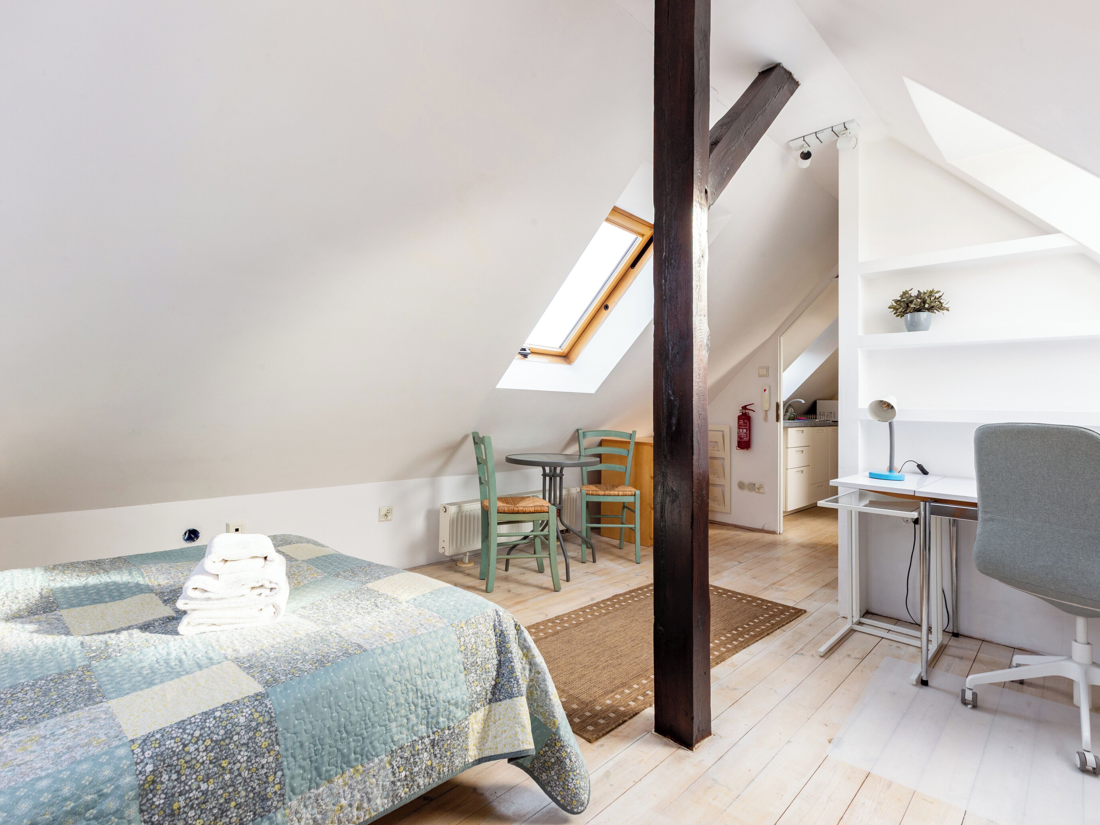 Une chambre mansardée chaleureuse, avec un lit, un bureau et une chaise. On y trouve une lucarne, un radiateur et une petite plante sur le bureau.