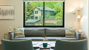 A room with a sofa facing a window overlooking a house, a small table with a decorative item, and a wall-mounted light fixture. - Walk to Main Street & Town Lift, Private Hot Tub, Garage Parking, On Shuttle Route (Park City)