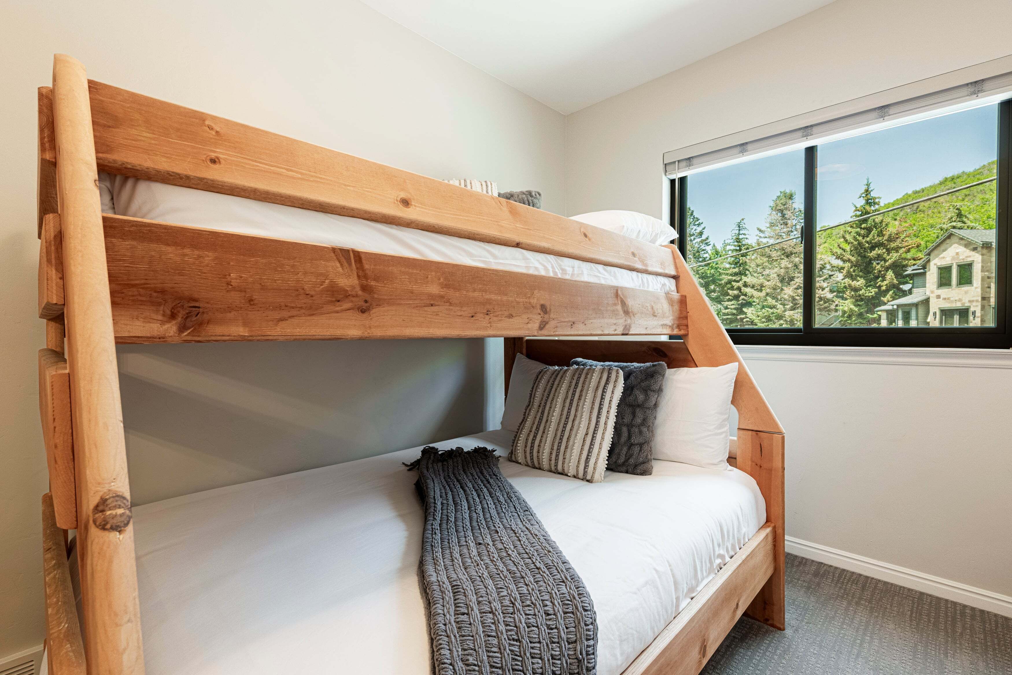A bunk bed with a wooden frame, white bedding, and a view of a house through a window.