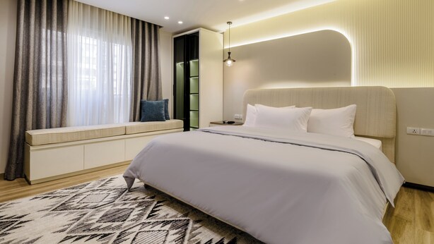 A modern bedroom with a large bed, a bedside table, a built-in bench with storage, and a large window with curtains. - 3 bedroom apartment (Giza Governorate)
