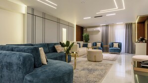 A modern living room with a sectional sofa, a round ottoman, and a coffee table. There is a dining area with a table and chairs, and a kitchen area with a refrigerator and a sink. - 3 bedroom apartment (Giza Governorate)
