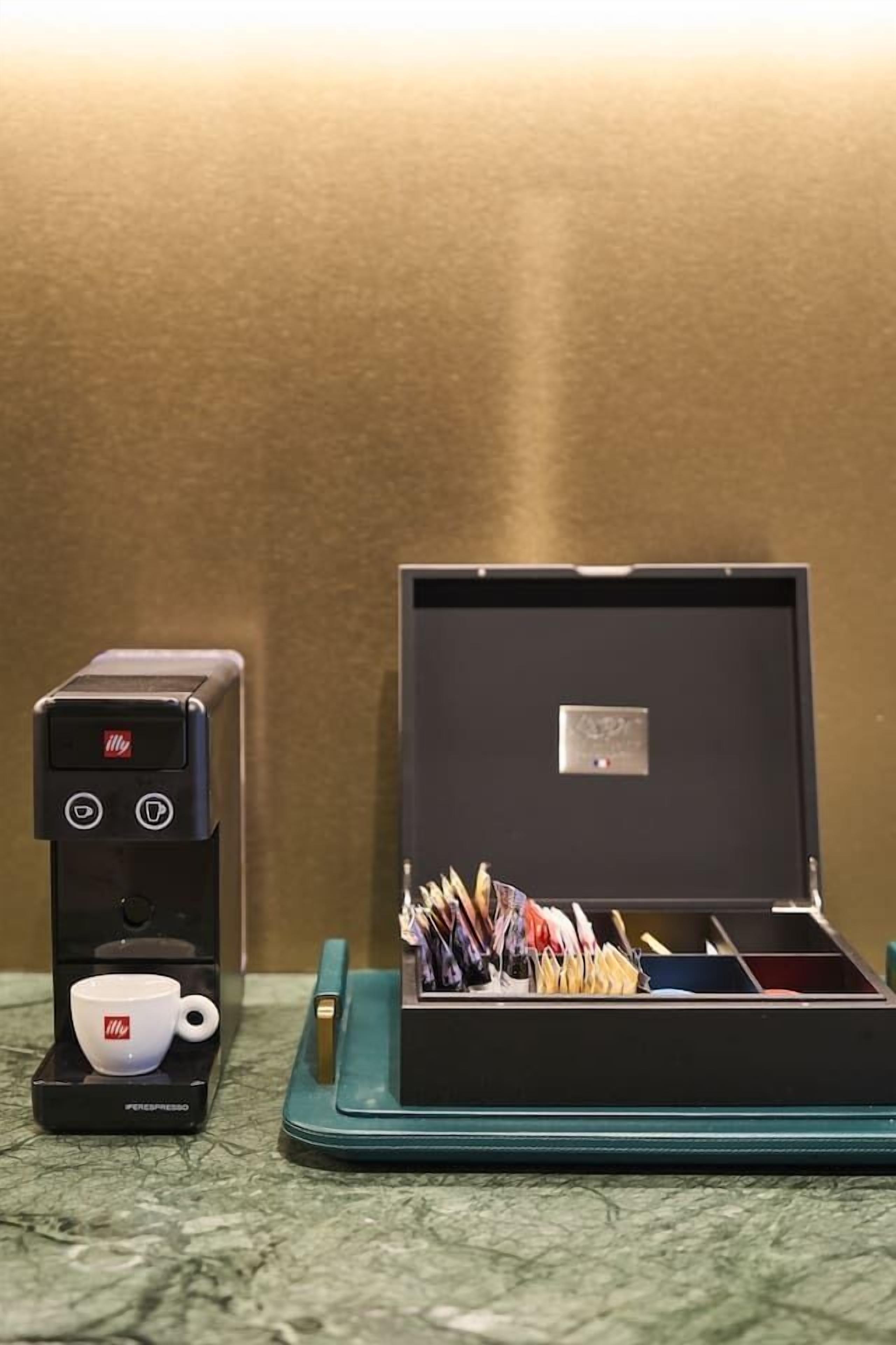 Une machine à café avec une tasse posée sur un plateau, un plateau turquoise sur lequel se trouve une boîte de stylos, et un mur en arrière-plan.