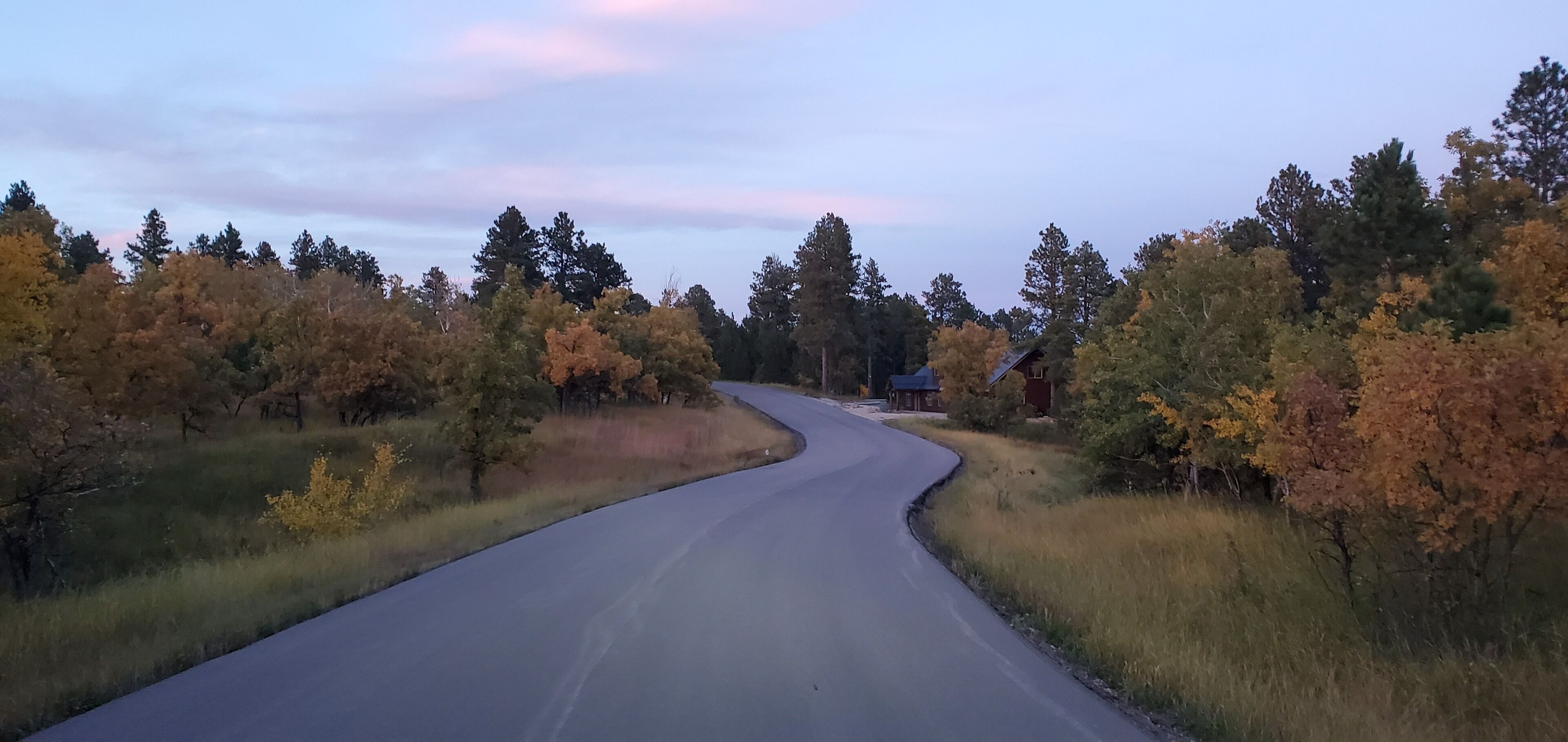 Mutkitteleva tie, jonka molemmin puolin kasvaa puita, joiden lehvistö on syksyn väreissä, johtaa pieneen taloon, jonka katto on punainen.