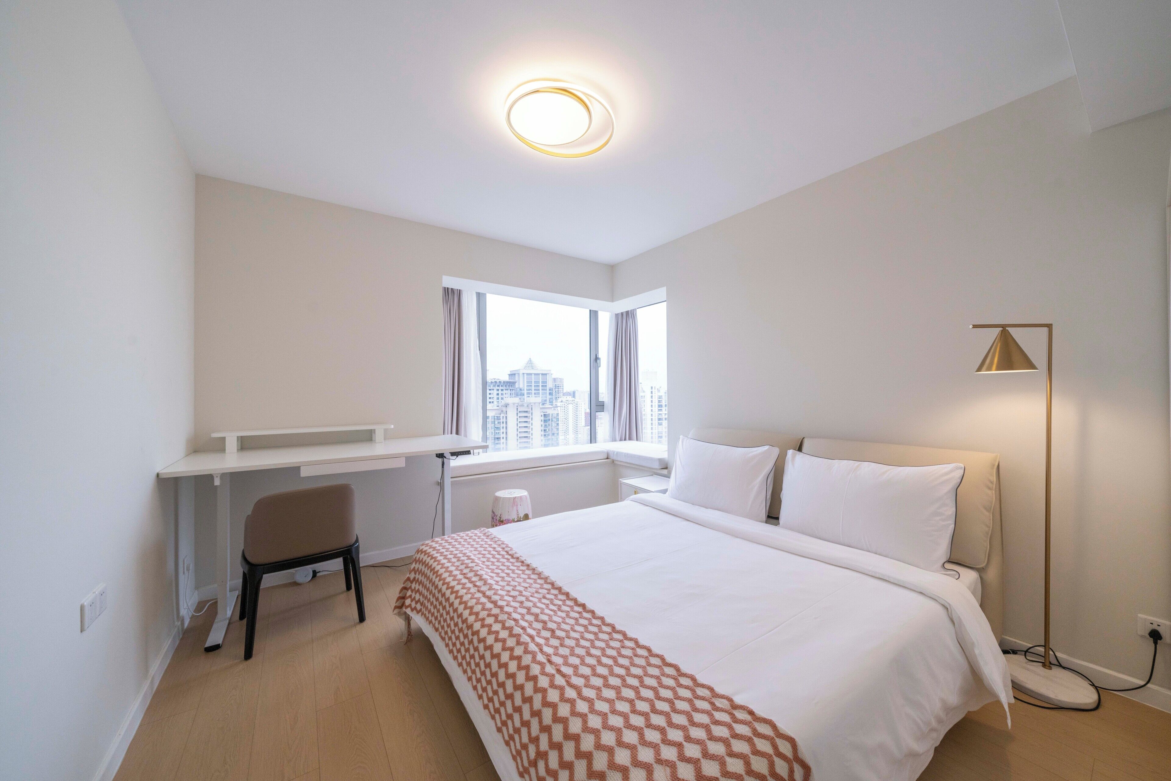 A modern hotel room with a large bed, a desk, and a chair. There is a window with a view of the city.