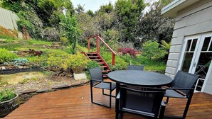 Une terrasse en bois avec une table ronde et des chaises, entourée d’une végétation luxuriante et d’un escalier rouge.