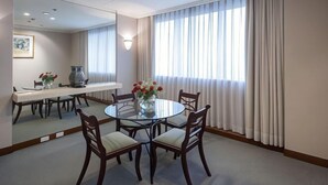 Un coin repas comprenant une table ronde en verre, six chaises, un vase avec des fleurs, un autre vase posé sur la table, un miroir au mur et une fenêtre avec des rideaux.