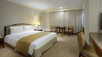 Quarto de hotel com uma cama grande, uma escrivaninha, duas cadeiras, uma televisão e uma janela com cortinas.