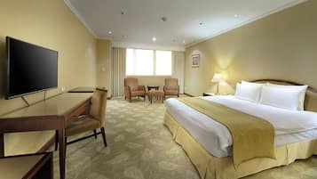 Quarto de hotel com cama, secretária, televisão e duas cadeiras.