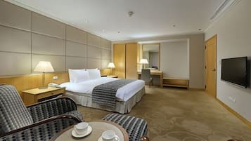Quarto de hotel com cama, escrivaninha, televisão e área de estar com duas cadeiras e uma mesa.