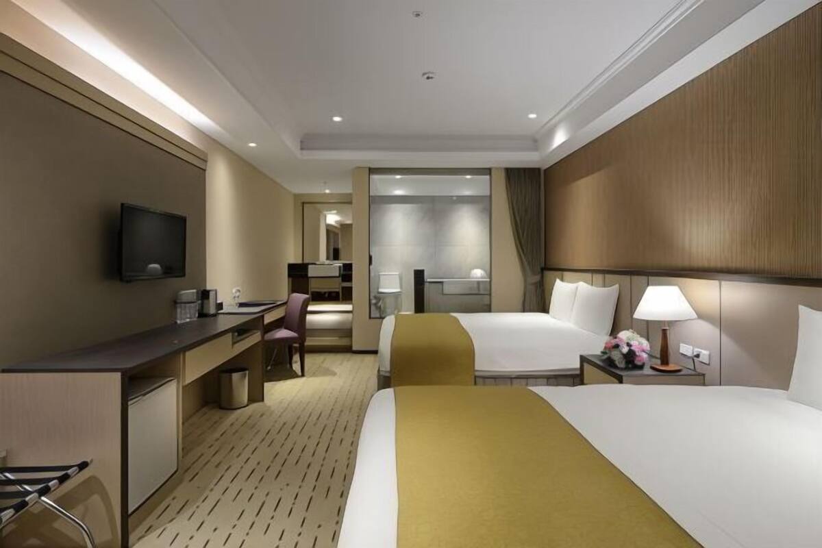 Habitación de hotel con dos camas, un escritorio, un televisor y un baño visible a través de una puerta abierta.
