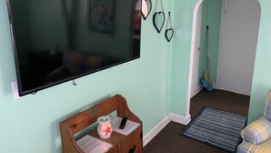 Uma sala de estar com uma televisão de tela plana na parede, um banco de madeira com uma vela e controle remoto, uma porta que dá para outro cômodo e um tapete estampado no chão.