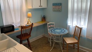Uma cozinha com micro-ondas, pia, duas cadeiras de madeira, uma mesa pequena com tampo azul e branco estampado, um armário branco e um aviso na parede.