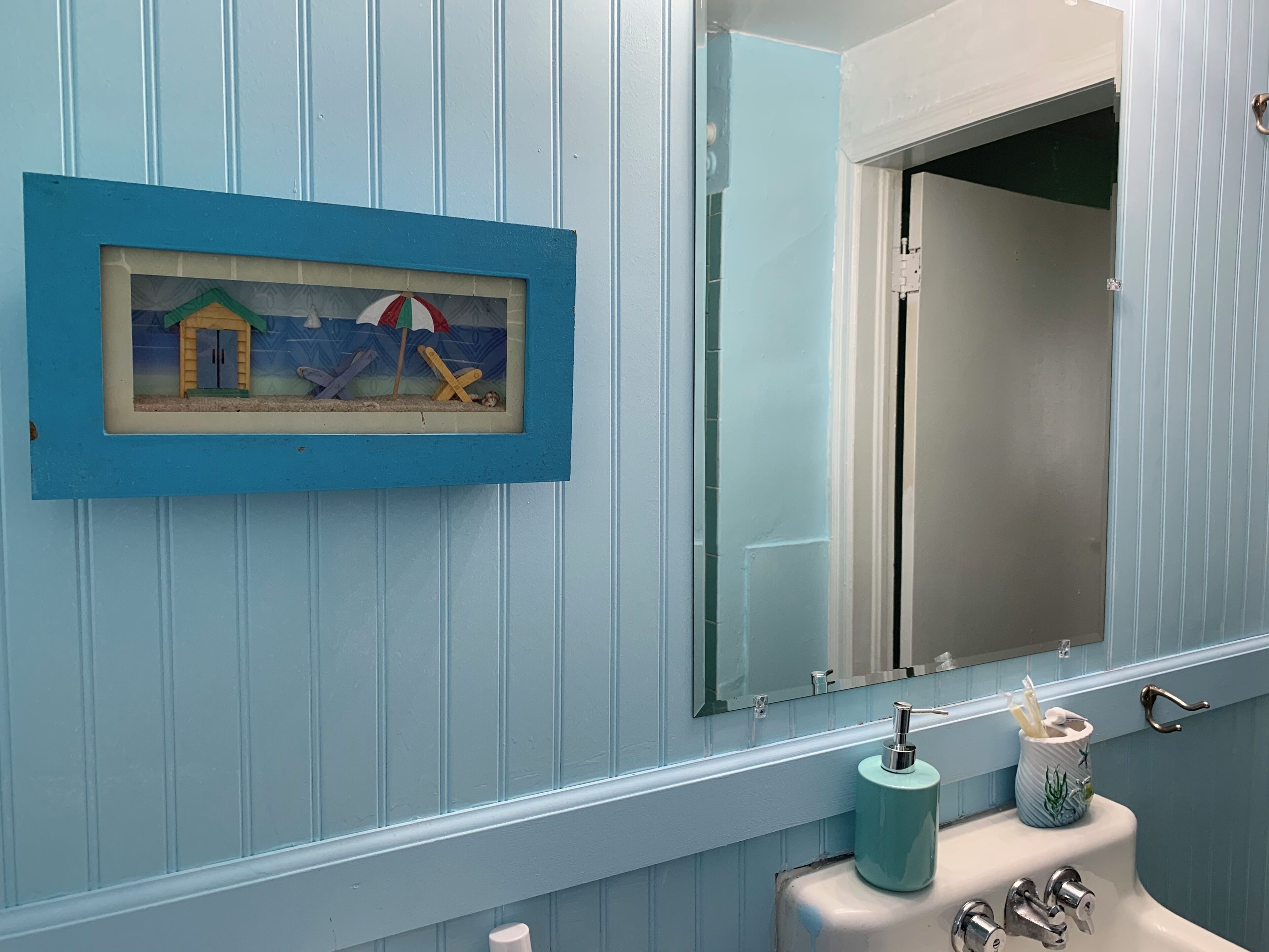 Un baño con pared azul, un cuadro enmarcado de una escena de playa, un lavamanos con dispensador de jabón y un espejo.