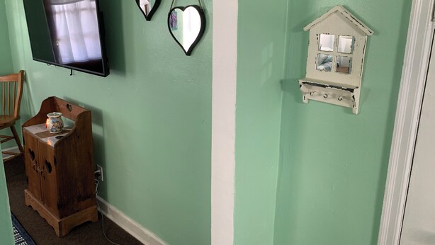 Quarto com televisão na parede, um armário de madeira com um vaso e uma pequena decoração branca em formato de casa.
