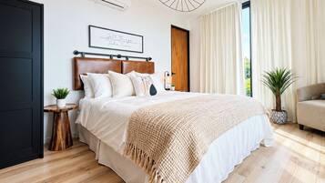 Un dormitorio moderno con una cama grande, cabecera de madera, mesita de noche con una planta y un ventanal con cortinas.