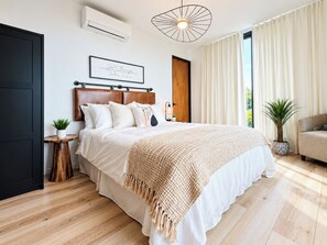 A modern bedroom with a large bed, a wooden headboard, a bedside table with a plant, and a large window with curtains. - Campo 5 Retreat W/ Private Climatized Pool (Coamo)