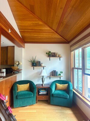 A room with wooden ceiling, two green armchairs, a small table with a lamp, and shelves with plants and books.