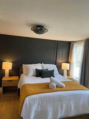 Une chambre d’hôtel avec un grand lit, deux tables de chevet avec des lampes, un mur de couleur sombre, une fenêtre avec des rideaux et un luminaire au plafond.