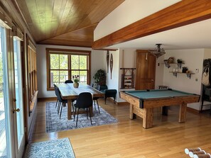 A spacious room with a pool table, wooden ceiling beams, a dining table with chairs, and a large window offering a view of greenery.