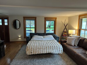A bedroom with a bed, a brown leather sofa, a standing lamp, and two windows with a view of the outdoors.