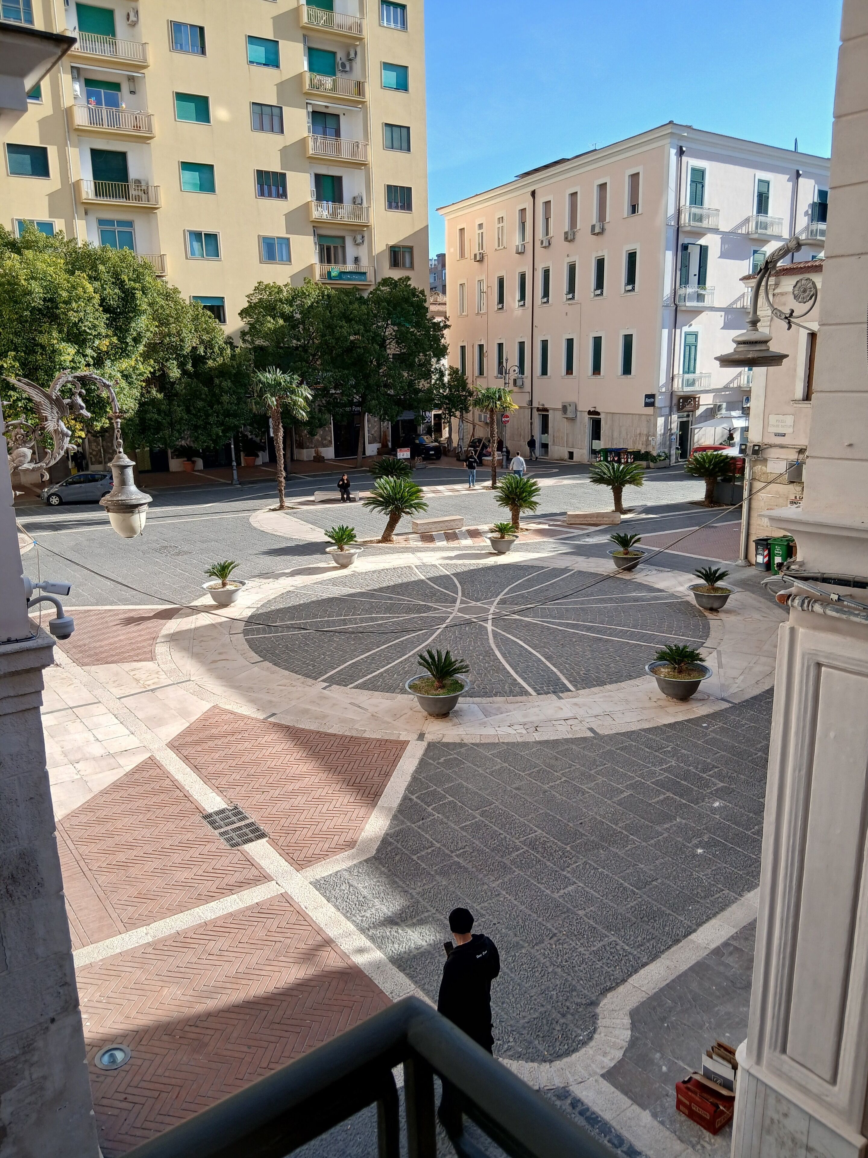 A courtyard with a central circular pattern, surrounded by potted plants and buildings.