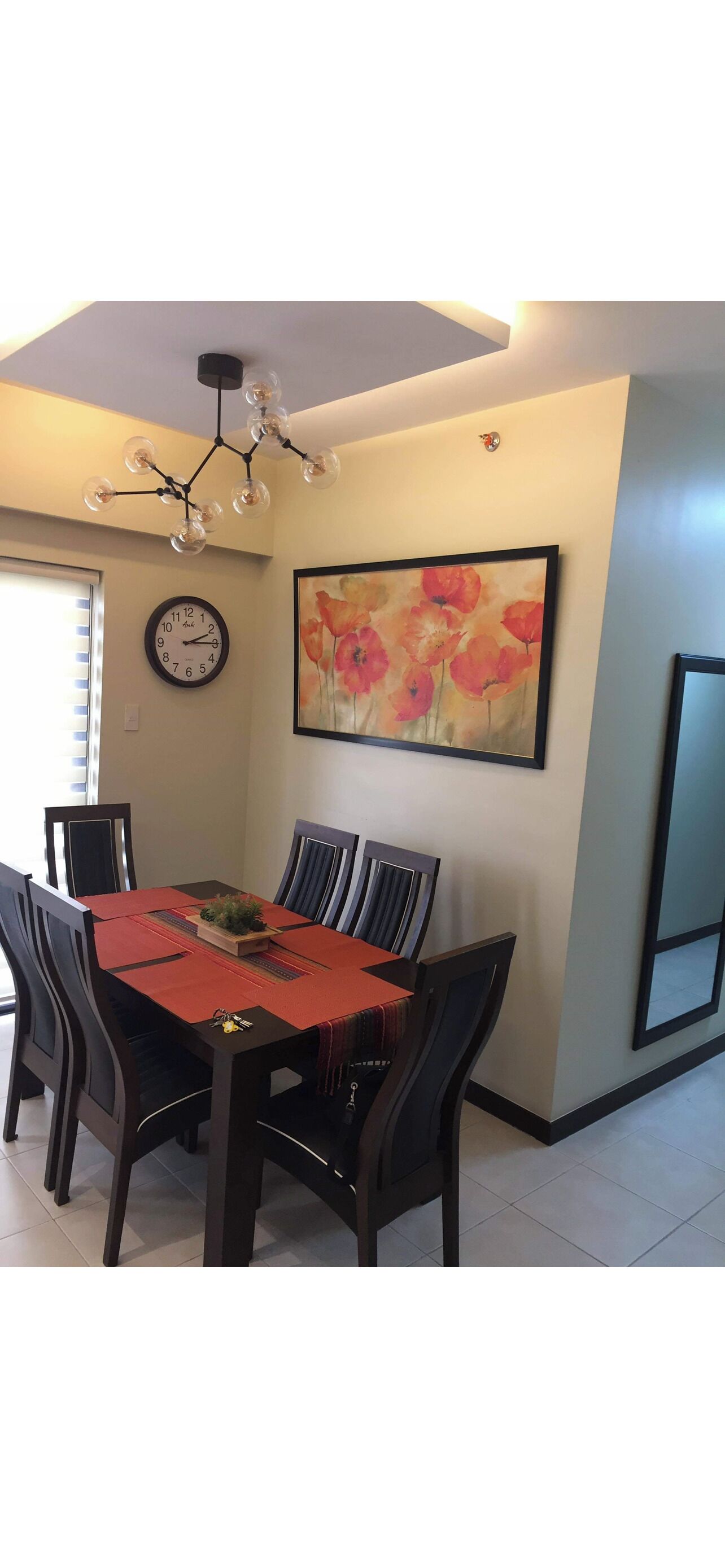 Un comedor con una mesa dispuesta para cuatro personas, rodeada de sillas, un reloj y un cuadro floral en la pared.