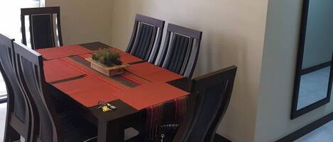 Un comedor con una mesa dispuesta para cuatro personas, rodeada de sillas, un reloj y un cuadro floral en la pared.