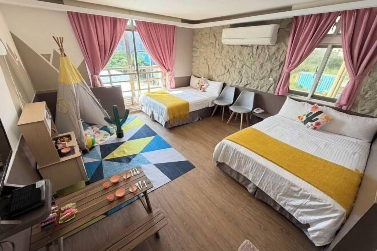 A hotel room with a bed, a desk with a laptop, a chair, a small table with items, and a window with curtains.