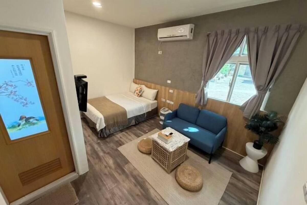 A room with a bed, a blue sofa, a small table, a plant, and an air conditioning unit.