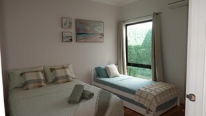 Une chambre avec un grand lit, un plus petit lit et une fenêtre avec des rideaux. Trois tableaux encadrés ornent le mur.