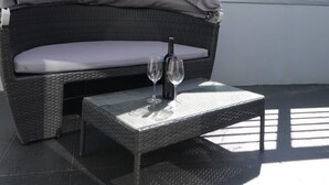 Un espace extérieur aménagé avec des meubles en osier, comprenant un banc rembourré et une table, ainsi qu’une bouteille de vin et deux verres.