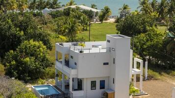 A modern two-story building with a pool, surrounded by greenery and a view of the sea.