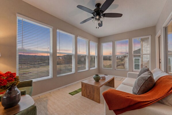 A spacious living room with large windows, a ceiling fan, and a view of the sunset.
