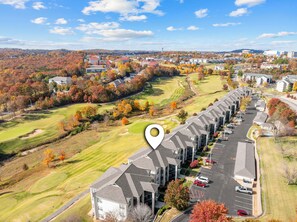 An aerial view of a golf course with a row of modern buildings, likely a hotel, with parking and a golf cart path.