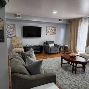 Ein Wohnzimmer mit grauem Sofa, grauem Sessel, Couchtisch aus Holz und einem flachbildfernseher an der Wand.