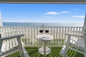 Un balcone con due sedie bianche e un tavolino con due bicchieri da vino e una rivista. Il balcone si affaccia su una spiaggia e l'oceano.