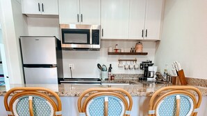 Cucina con mobili bianchi, frigorifero in acciaio inossidabile e microonde. Tre sedie in vimini con cuscini blu e finiture bianche. Un tappeto colorato è sul pavimento.