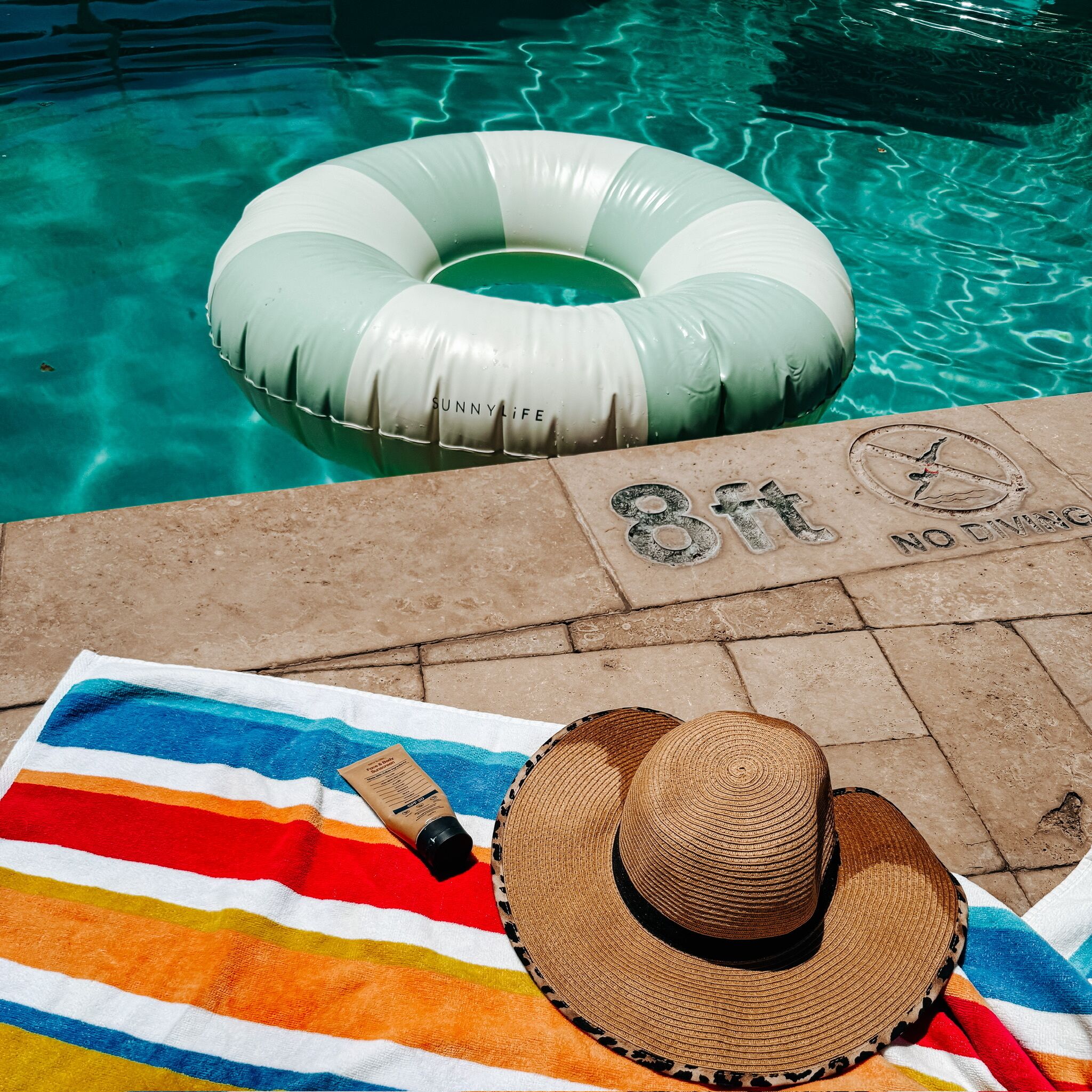 Área da piscina com um flutuador, uma toalha listrada, chapéu e frasco de protetor solar.