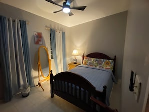 Ein Schlafzimmer mit einem Bett, einem Deckenventilator, einem Nachttisch mit Lampe, einem Spiegel und einem Fenster mit Vorhängen.