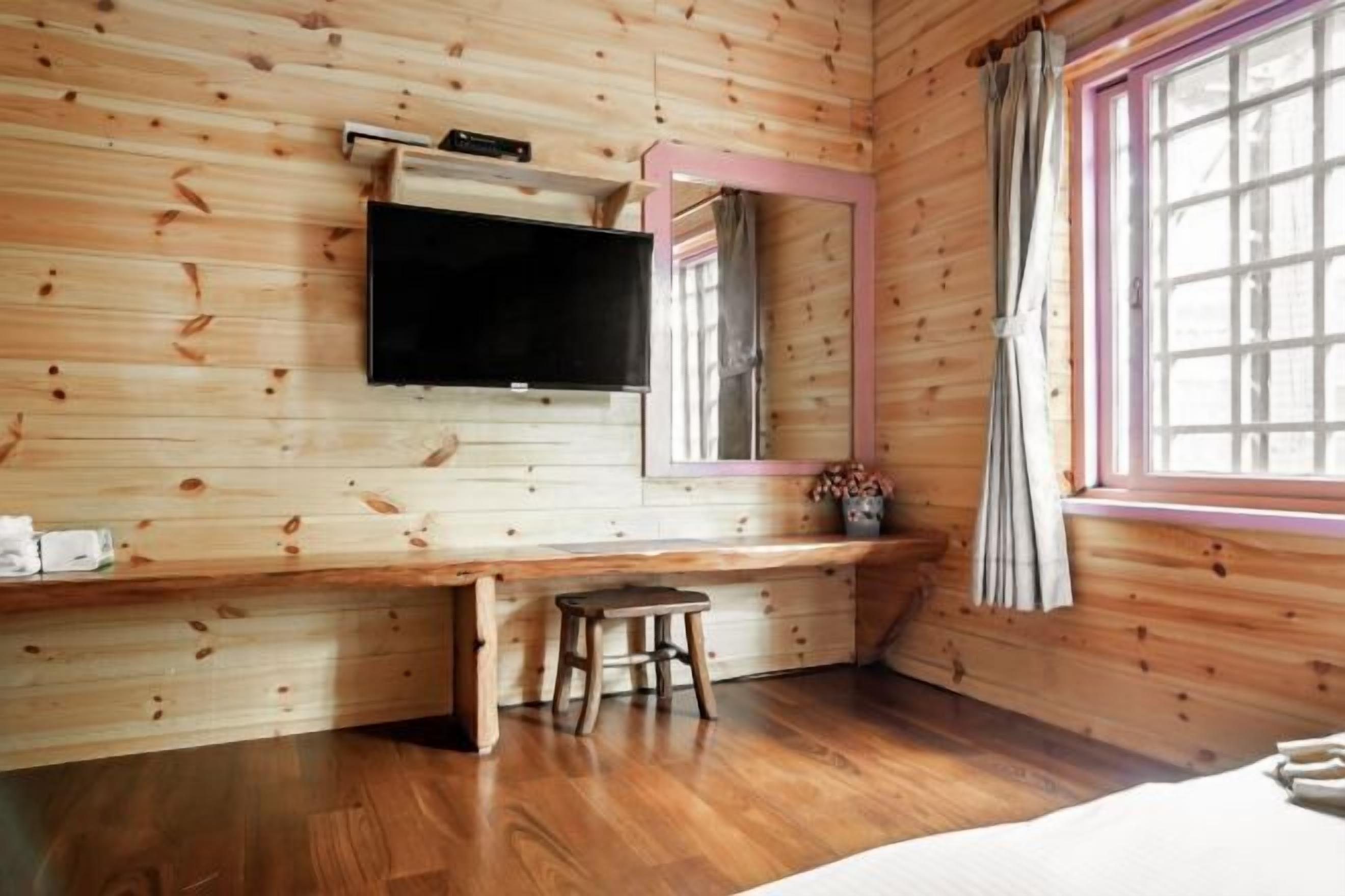 A wooden cabin room with a bed, a desk with a chair, a TV mounted on the wall, and a window with curtains.