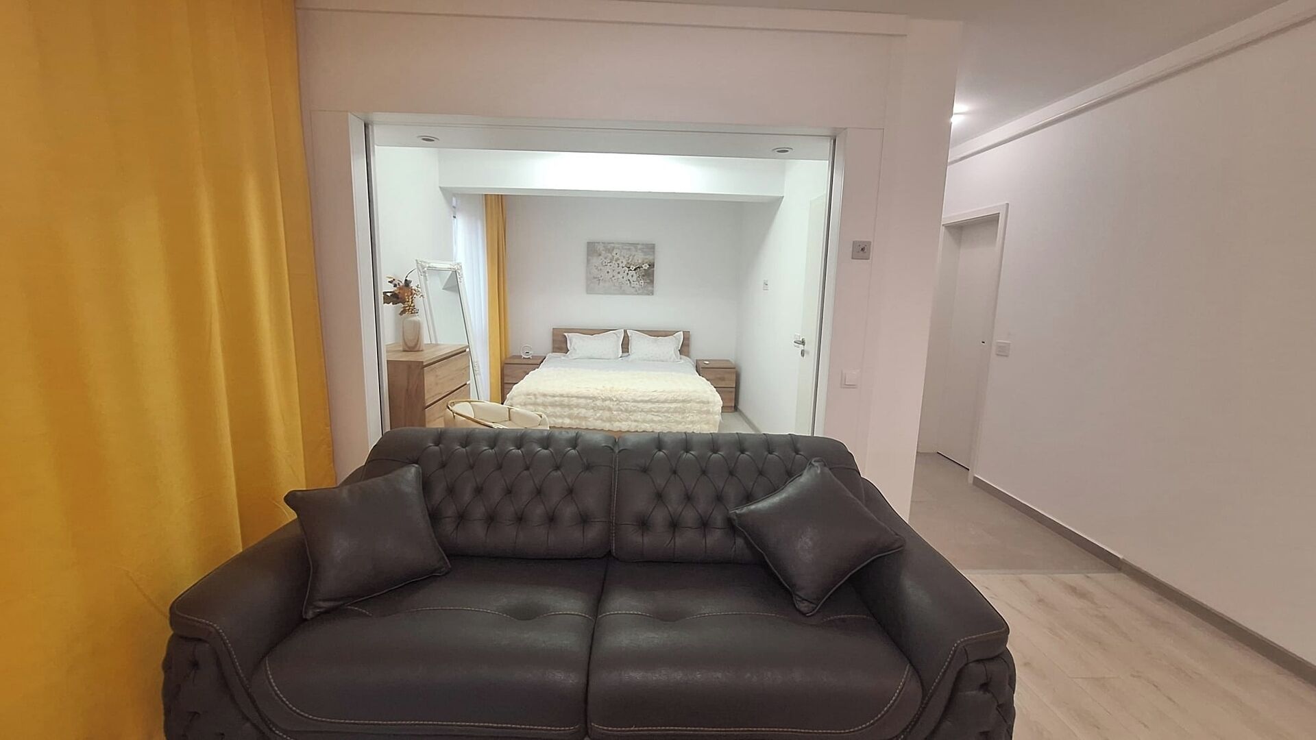 Un dormitorio moderno con un sofá de cuero oscuro, una cama con ropa blanca, una mesita de noche y un espejo.