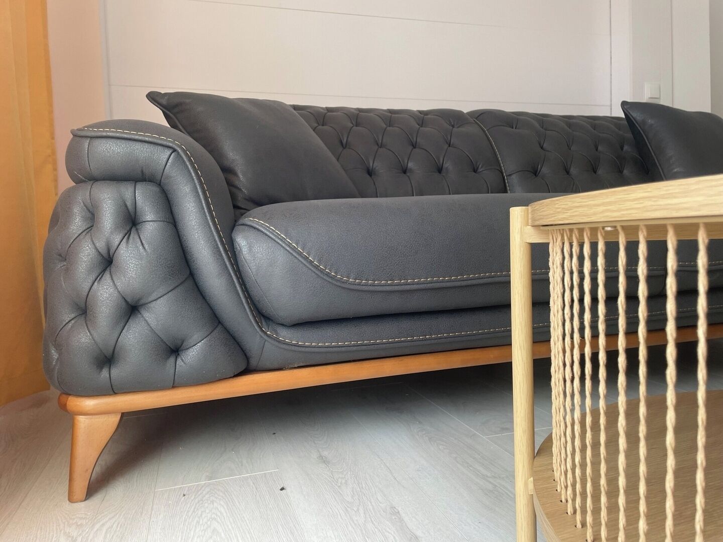 A dark grey tufted sofa with wooden legs and a wooden side table with ropes.