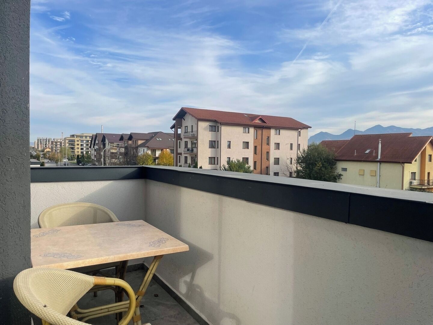 Een balkon met een tafel en twee stoelen, met uitzicht op een woongebied met gebouwen en bergen op de achtergrond.
