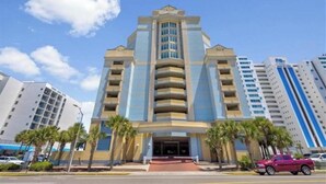 A multi-story hotel with a central entrance, surrounded by palm trees and other buildings. - Stunning 10th-Floor 1-Bedroom Oceanview Condo (Myrtle Beach)
