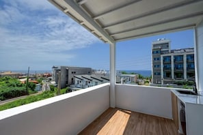 Un balcon doté d’un plancher en bois, de murs blancs et offrant une vue sur des bâtiments et de la verdure.