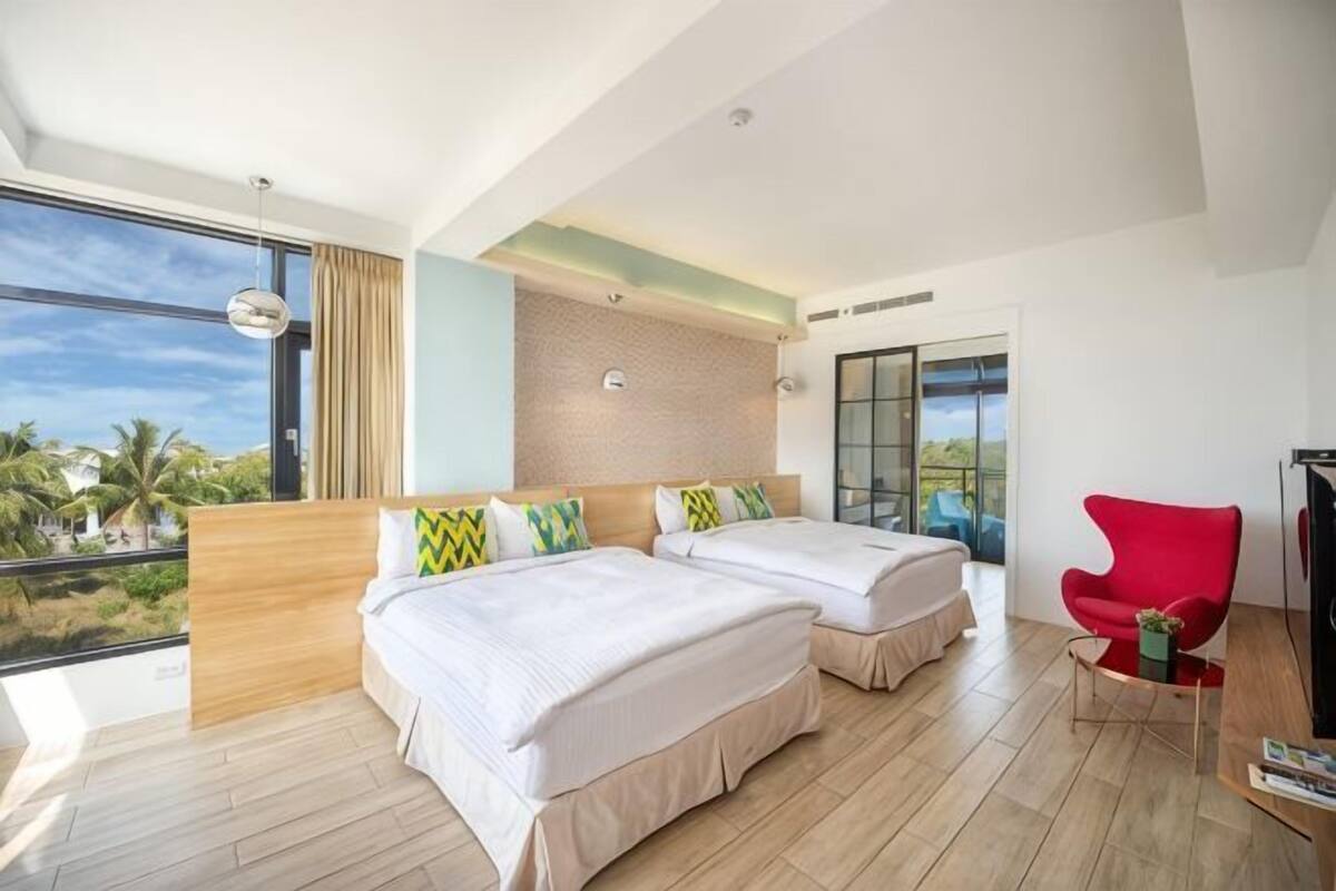 A hotel room with two beds, a red chair, a TV, and a large window with a view.