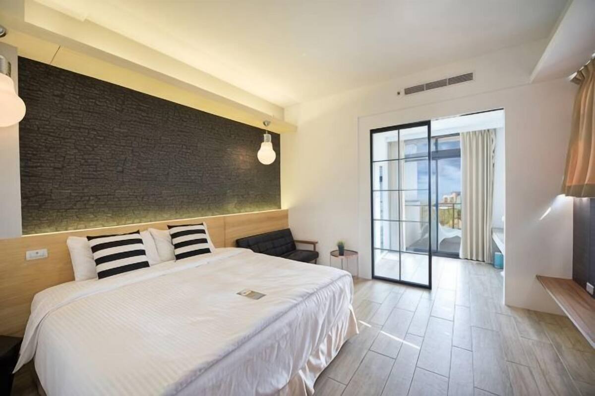 A modern hotel room with a large bed, a dark textured wall, a glass door leading to a balcony, and a wooden bench.