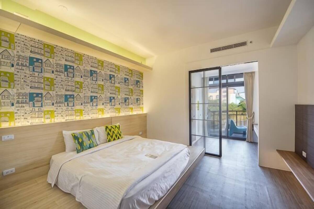 A modern hotel room with a large bed, a cityscape mural, a balcony with a view, and a flat-screen TV.
