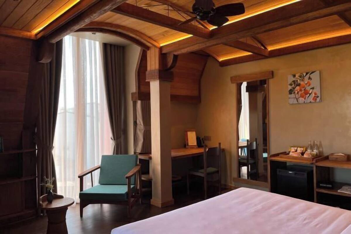 A hotel room with a large bed, a desk, a chair, and a small table. There is a window with curtains, a ceiling fan, and a painting on the wall.