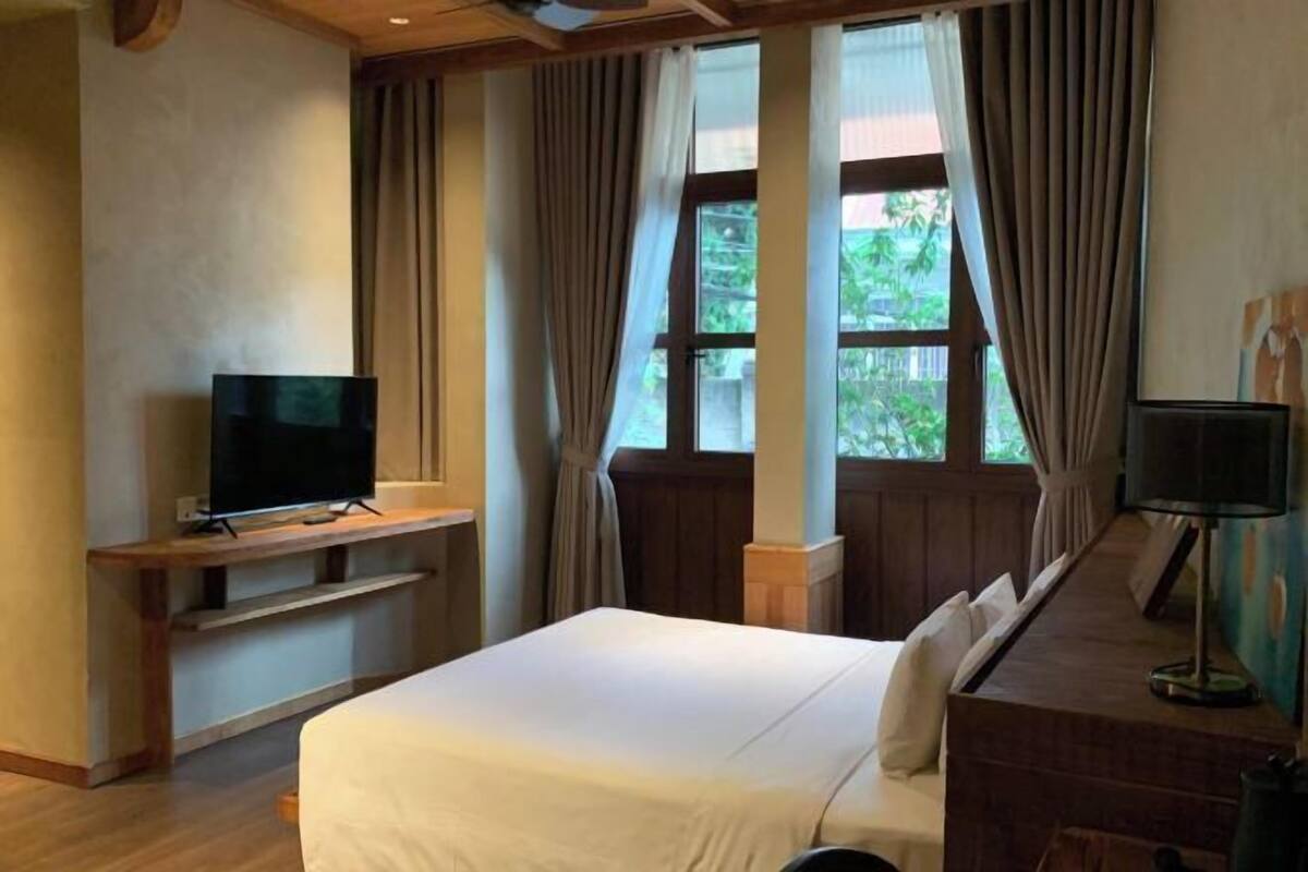 A hotel room with a bed, a TV on a wooden stand, a desk with a lamp, and large windows with curtains.